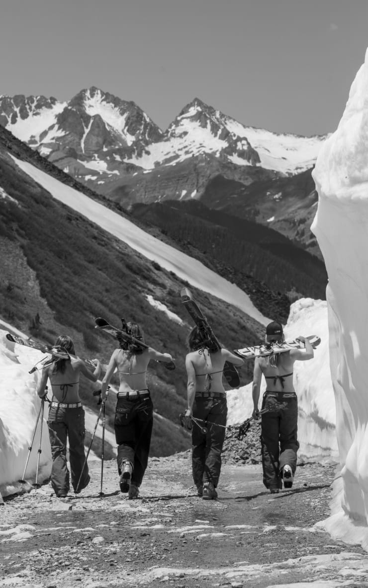 Black and white photo of four girls with bikini tops holding skis walking toward a mountain by Slate Gray Gallery Artist Brett Schreckengost