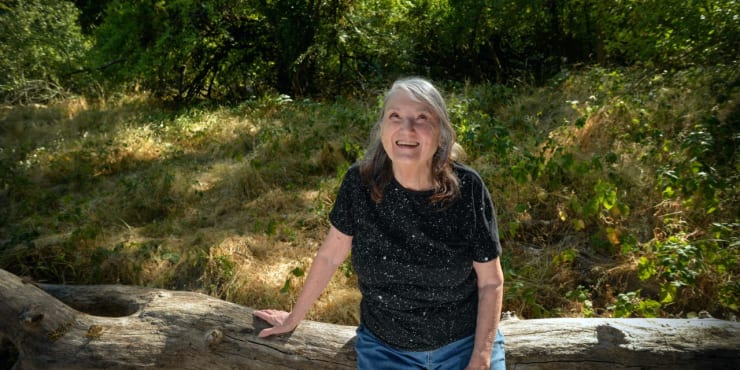 (Above: Margaret Coe sits on a fallen tree trunk in the meadow behind her home. The “au naturel” area provided the inspiration for her latest art exhibit, Wild Spaces, at the Karin Clarke Gallery; photos by Paul Carter.)