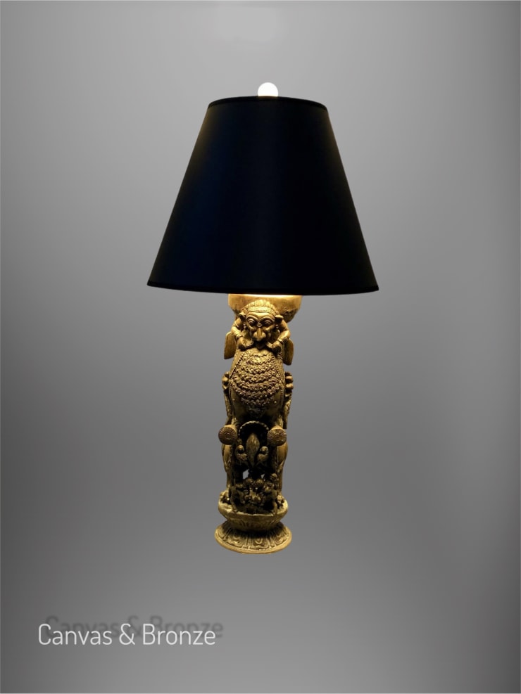 A decorative lamp featuring an ornate, golden statue base with intricate detailing, topped by a sleek black lampshade against a plain background.