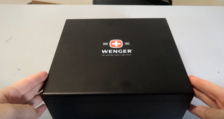 Hands opening a black box labeled "Wenger" on a table.