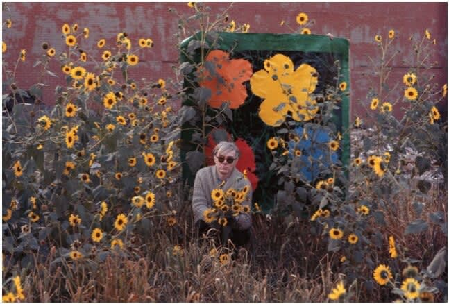 Andy Warhol surrounded by flowers in front of Flower with an early Flowers canvas, Queens, New York, 1964. ©2010 William John Kennedy, ©2012 Andy Warhol Foundation / Artists Rights Society (ARS), New York.
