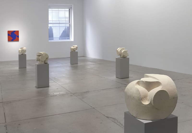 8 small white stone sculptures on pedestals in front of a red abstract painting on a white wall.