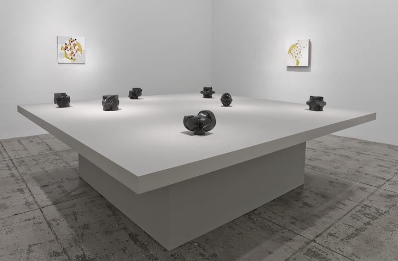 7 small stone sculptures on a table in front of two abstract paintings.