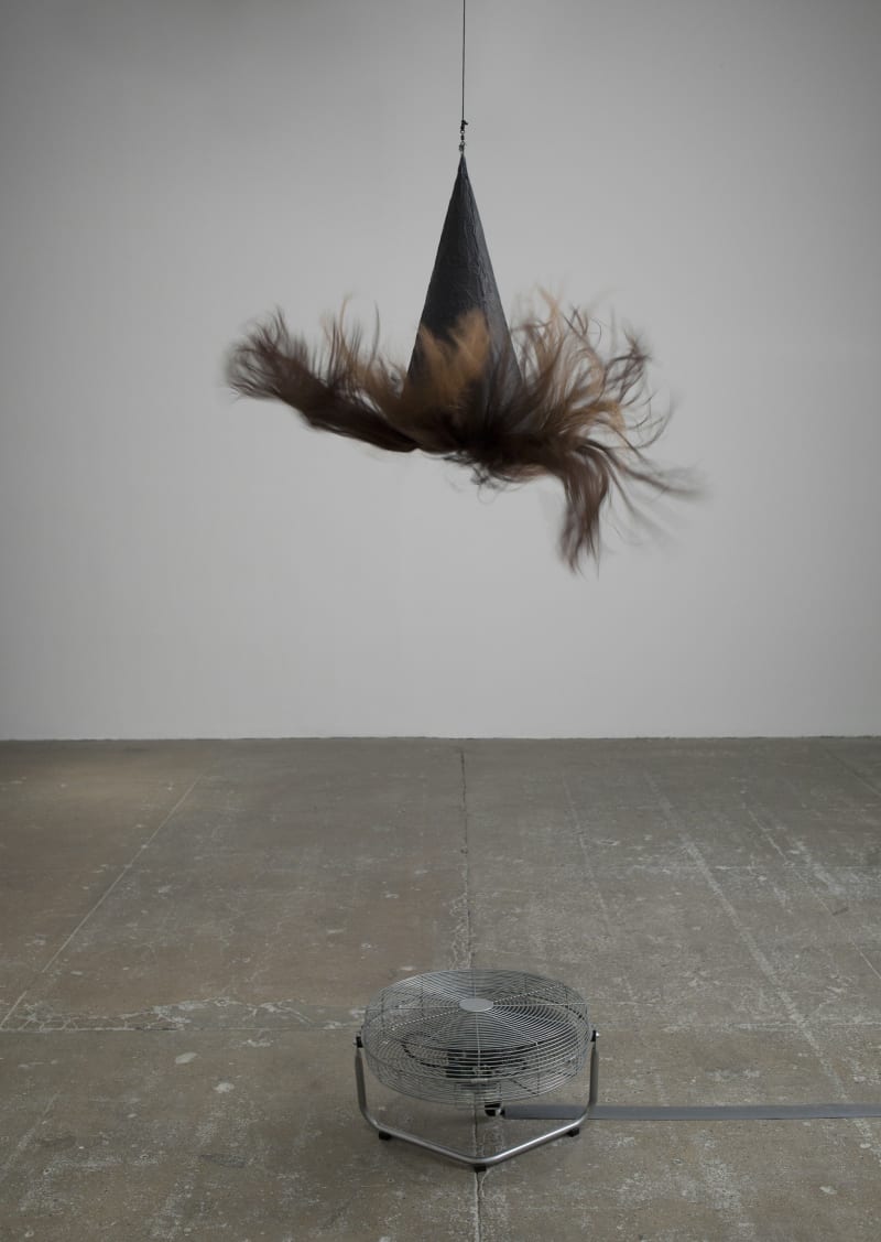 A witch's hat with long brown hair hangs suspended above a fan.