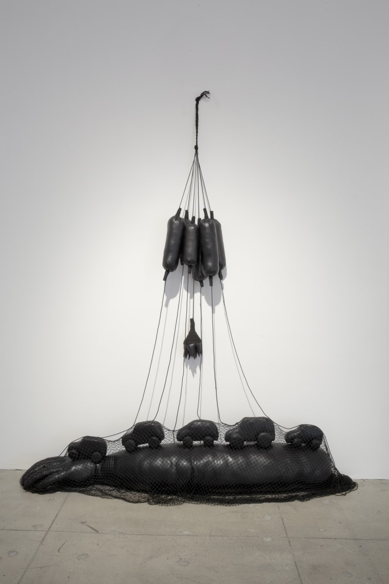 A black leather sculpture consisting of several small long balloons that is tied to a long leather arm on the floor that has cars driving on top of it.