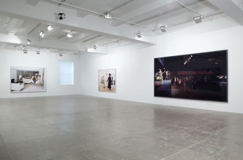 An image of two boys boxing, an image of an elegant woman dressed in black and an nighttime image of an outdoor concert are displayed in a white room. 
