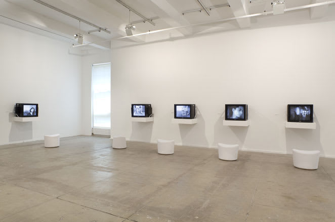 Gallery view of cathode ray televisions on shelves with stools underneath.