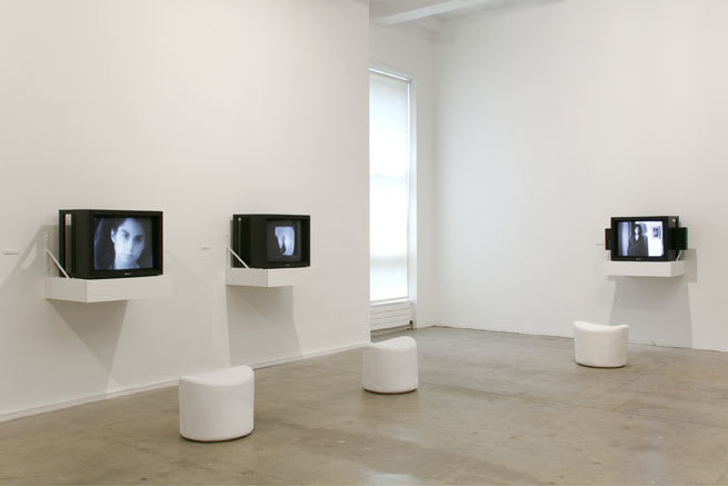 Gallery view of cathode ray televisions on shelves with stools underneath.