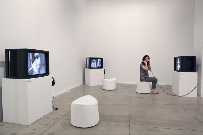 Gallery view of cathode ray televisions on shelves with stools underneath and one seated viewer.