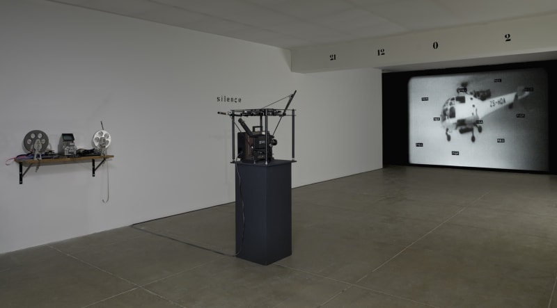 In a white room with a beige floor, a projector runs, showing a large image of a helicopter with small scattered text over it. A beam on the ceiling of the room reads: 21 12 0 2. On the left wall, the text reads: silence.