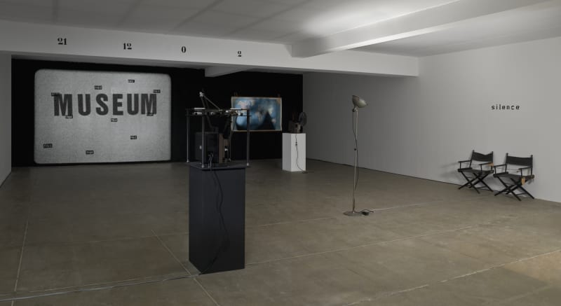 In a white room with a beige floor, two projectors run, showing a world map on one, and a large image of that says MUSEUM with small scattered text over it. A beam on the ceiling of the room reads: 21 12 0 2. On the right wall, text reads: silence.