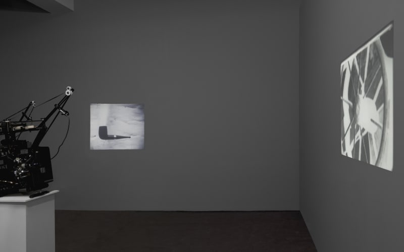 In a dark room, two projections play. Left: a pipe. Right: a wooden wheel.