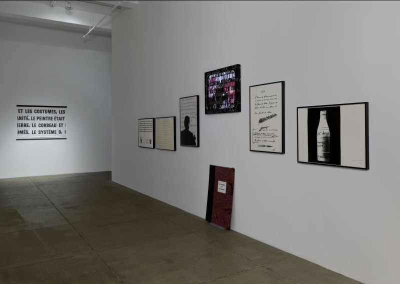 On the right white wall, various framed works of drawing and text hang. On the left, more texts hangs, reading: ET LES COSTUMES, LES ANITÉ, LE PEINTRE ÉTAIT IERRE, LE CORBEAU ET IMÉS, LE SYSTÈME D,
