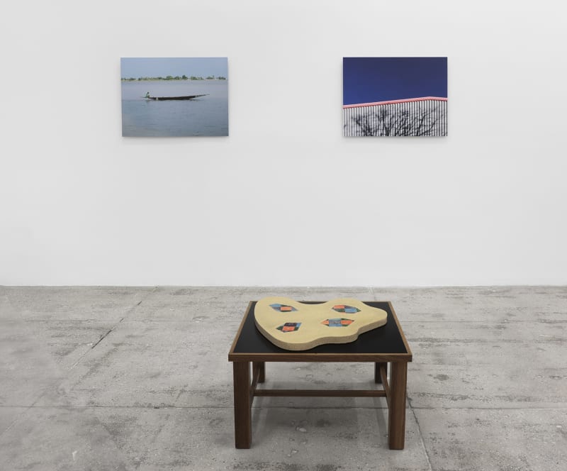 2 photographs hang on a white wall; on the left, a canoe in water and on the right, a roof and sky. A flat sculpture sits on a platform in the foreground. 