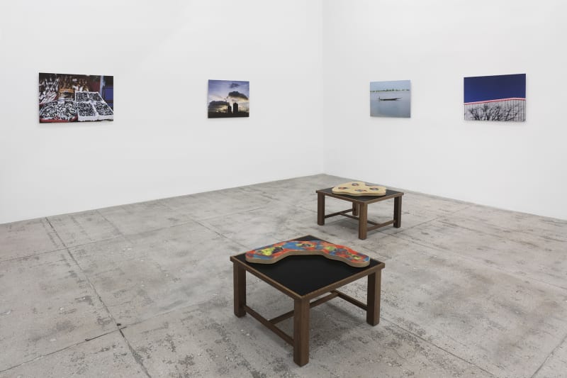 4 photographs hang in the corner of a large white room; 2 flat sculptures sit on wooden platforms in the foreground. 