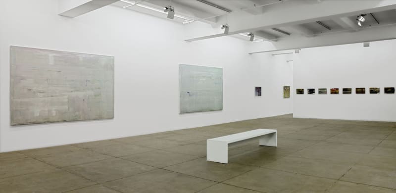 On the left, two large, gray abstract paintings hang on the wall. On the right is a row of 10 small, dark, abstract paintings. A white bench sits in the center of the room. 