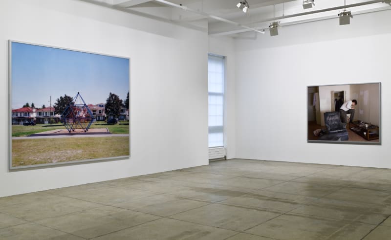 2 large, framed color photographs hang in the corner of a white room; on the left, a playground and on the right, a man in a dingy living room. There is a window in-between them.