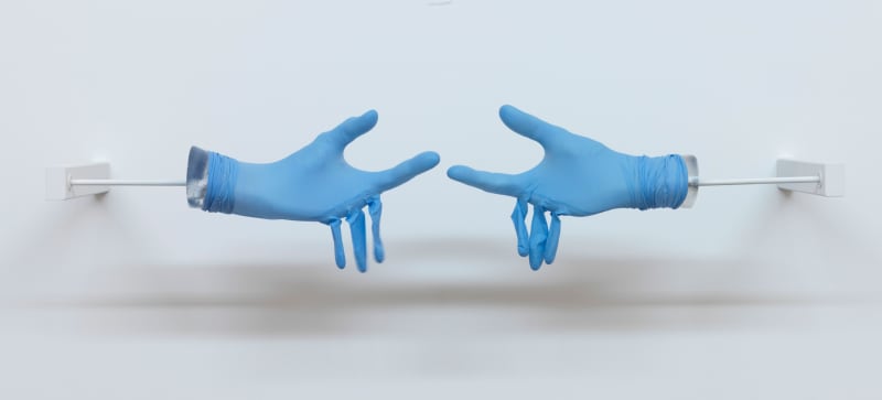 Two blue medical gloves are stretched over plaster hands reaching for each other. The middle, ring and pinky finger are empty and hang limply. 