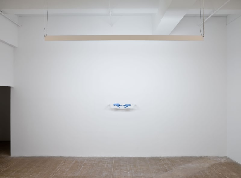 Two blue medical gloves stretched over plaster hands are seen pointing towards each other from a distance in front of a white wall. A fluorescent lamp hangs above them. 