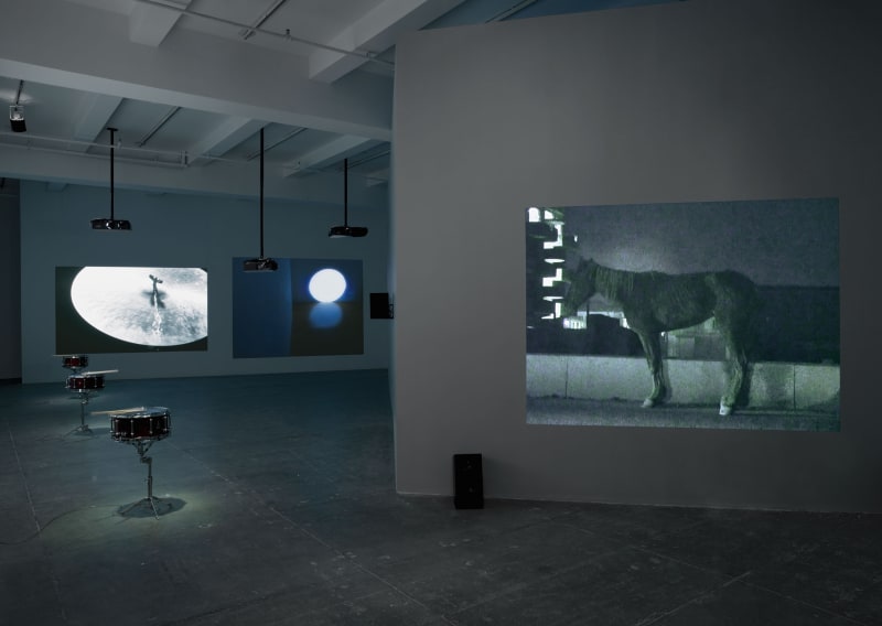 3 projections fill a darkened room while 3 snare kits sit in a line on the left. The projections display a cymbal, a circle of light, and a horse. 