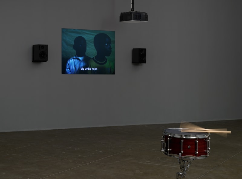 A snare-kit with moving drum sticks stands in front of a projection of two young black boys with the subtitle, big white hope. 