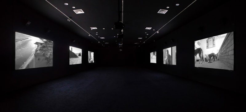 Six black and white images are projected on both sides of a dark room. 