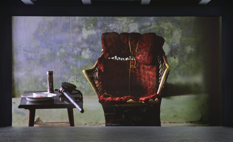A large projection depicts a wicker chair with a red cushion, a side-table with a rifle, two plates and a metallic bottle on top. 