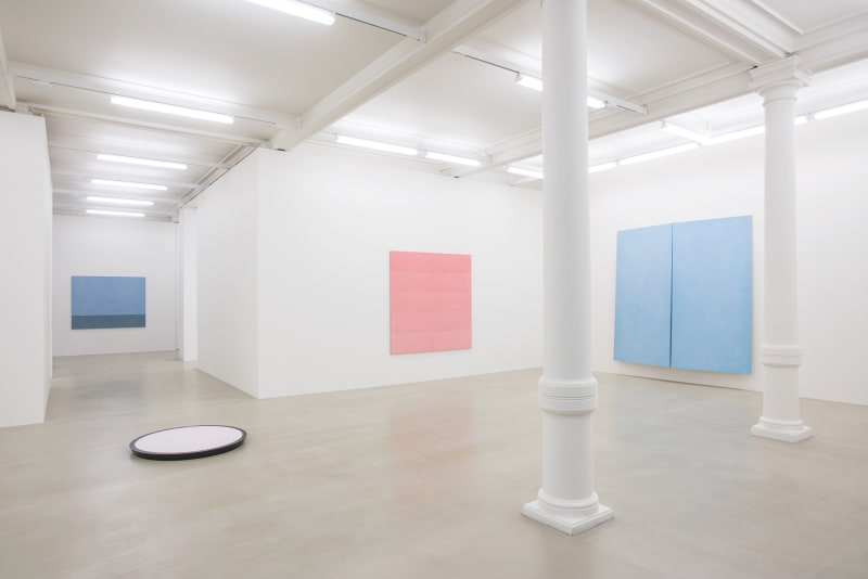 3 pastel paintings hang on the wall around a corner. There are 2 white columns in the room and a circular white object on the floor.