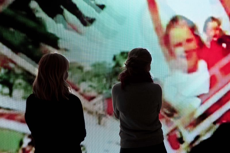 2 silhouetted figures look at a large projection of people on an amusement park ride. 