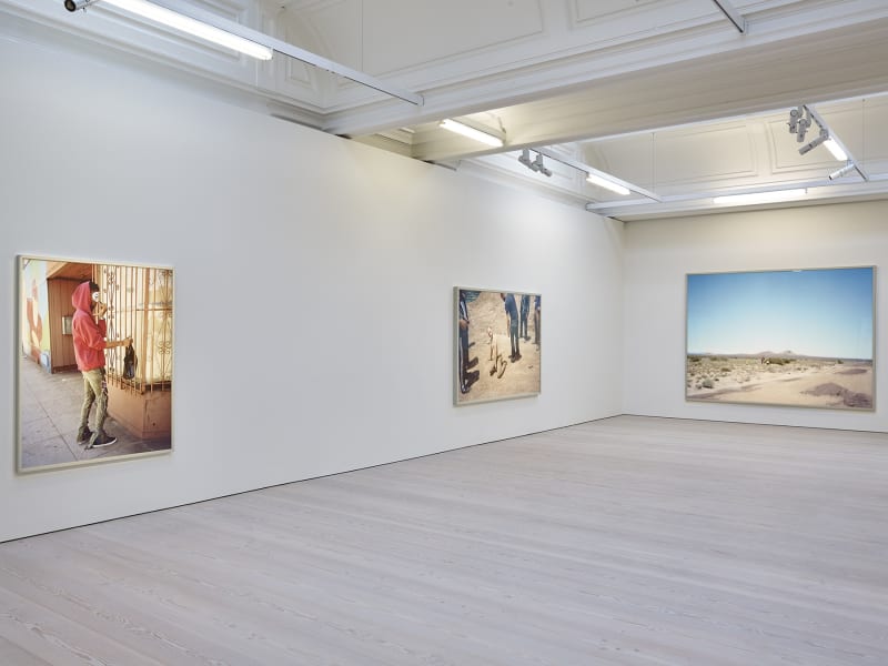 3 large photographs hang in a white room. From left to right: A man in a red sweatshirt and white mask; a shirtless man on the ground surrounded by 6 clothed men, and a desert landscape.