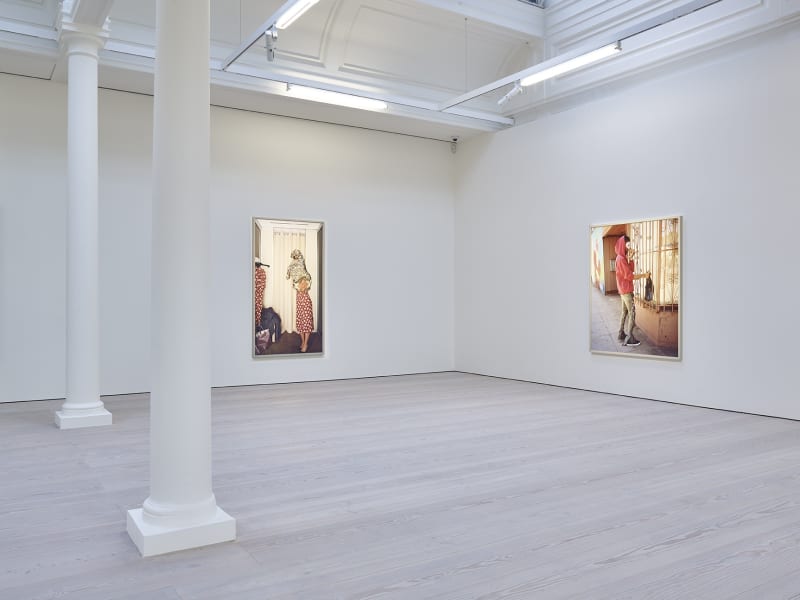 On the left hangs a photograph of a figure in a red polka dot dress; while on the right is a photograph of a man in a red sweatshirt and white mask. 