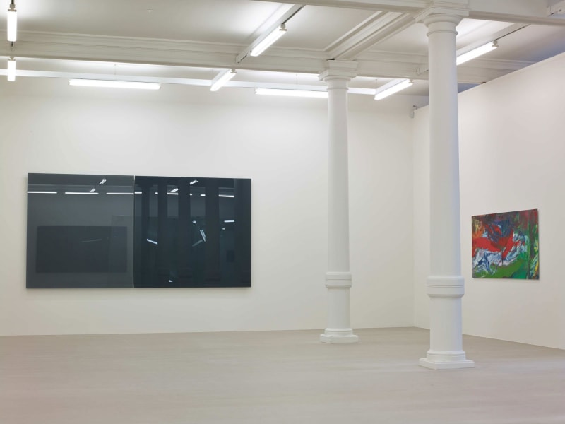 In a large white space with columns, two large square glass panes hang next to each other on a wall, one black and one grey. On the wall to their right is a small abstract painting, of reds, whites, and greens.