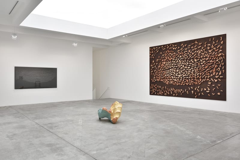 In a large white space with a skylight, a large brown canvas hangs on the right wall, covered in mounted wood objects. A wood sculpture sits on the floor in front of it, and a black painting hangs on the opposite wall.