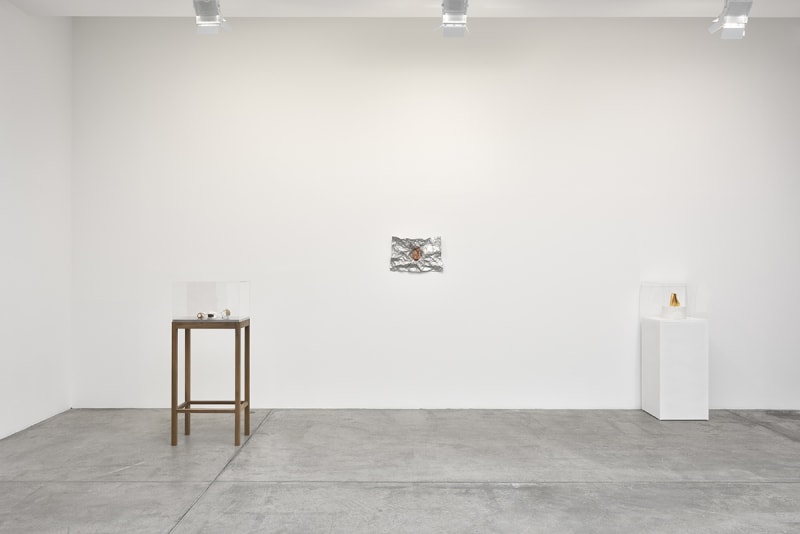 Between two elevated display cases with small objects, a small abstract painting hangs on the wall. It appears to be made up of crumpled tin foil.