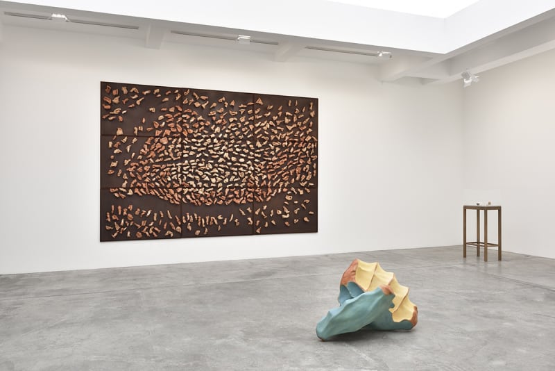 In a large white space with a skylight, a large brown canvas hangs on the wall, covered in mounted wood objects. A wood sculpture sits on the floor in front of the painting, and an elevated display case with small objects sits next to it.