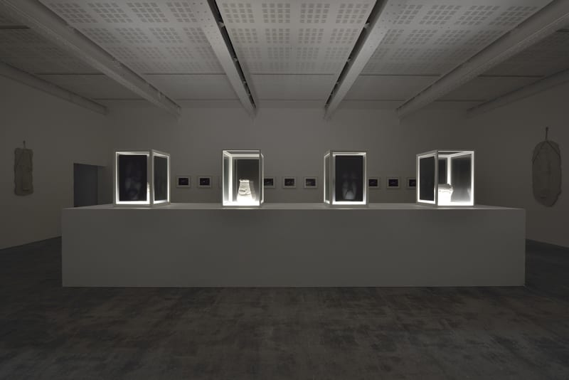 In a dark room, there are no lights except for those that illuminate, from below, white pottery in dark shaded display cases on a large white platform.