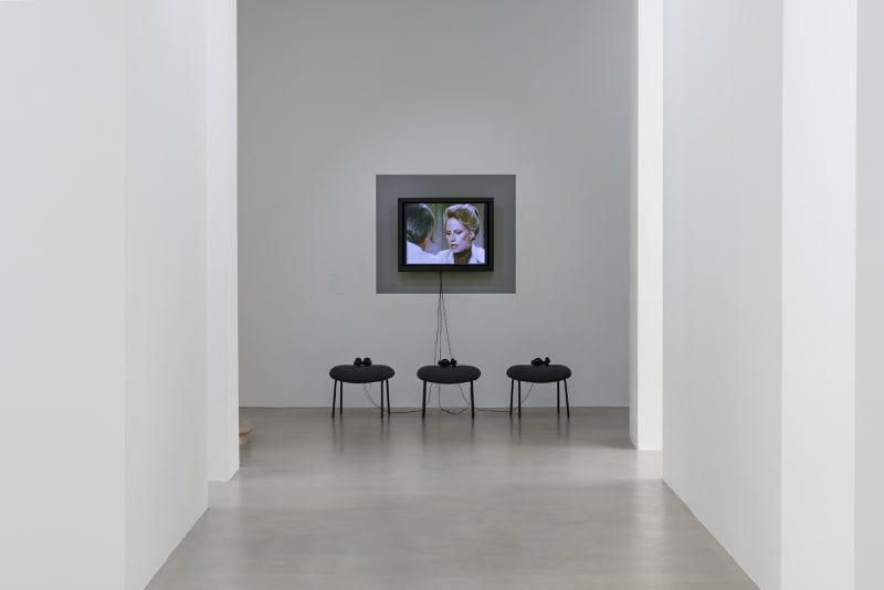 3 stools with headsets sit in front of a flatscreen monitor displaying a woman and a man conversing.