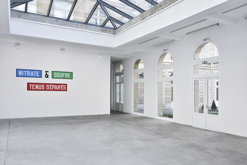 Multicolored text on the wall which reads "NITRATE & SOUFRE TENUS SEPARES". A row of windows is to the right.