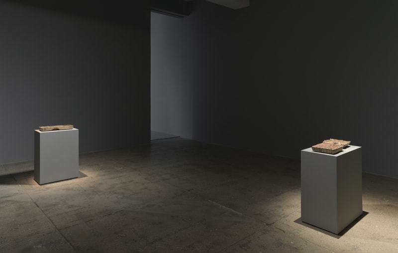 2 grey pedestals spotlit in a grey room displaying flat objects.