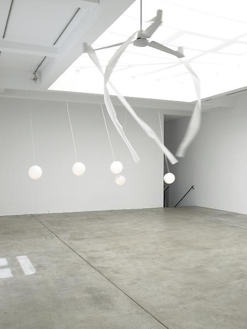 A gallery room with skylights has works hanging from the ceiling that move with the ceiling fan. 