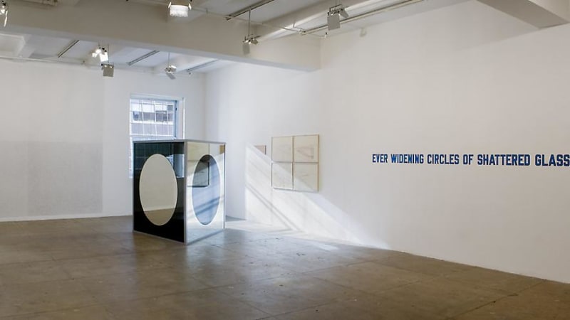 Light coming through the window on a glass sculpture. The text "EVER WIDENING CIRCLES OF SHATTERED GLASS" is on the wall.