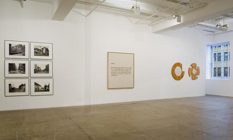 6 framed photographs, 1 framed painting and 2 wooden works hanging across a white wall. There is a window on the right. 