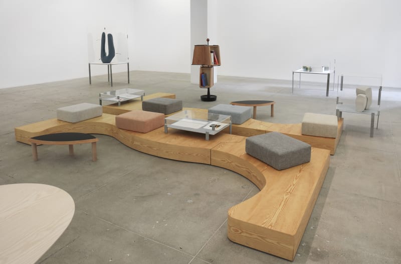An installation view of sculptures, custom furniture and cushions in a white gallery space.