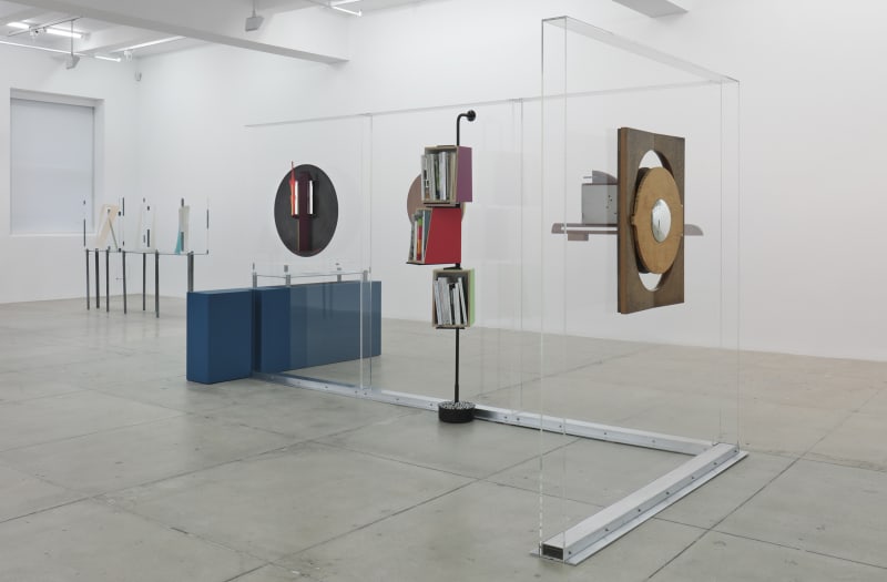 Various mirrors and a bookcase hang from a glass divider in the center of a white room.