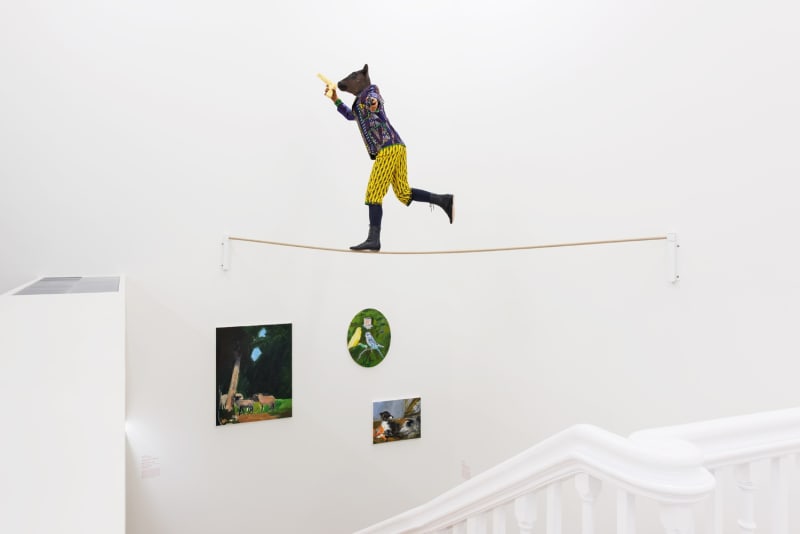 Installation view of miniature figure with a cow head and human body in wildly colored garments on a tightrope clutching a gun.