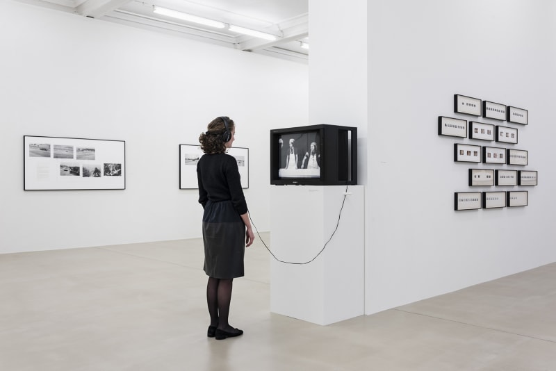 A person with headphones watches content on a cathode-ray tube tv displayed on a pedestal, surrounded by several black and white photographs on walls.
