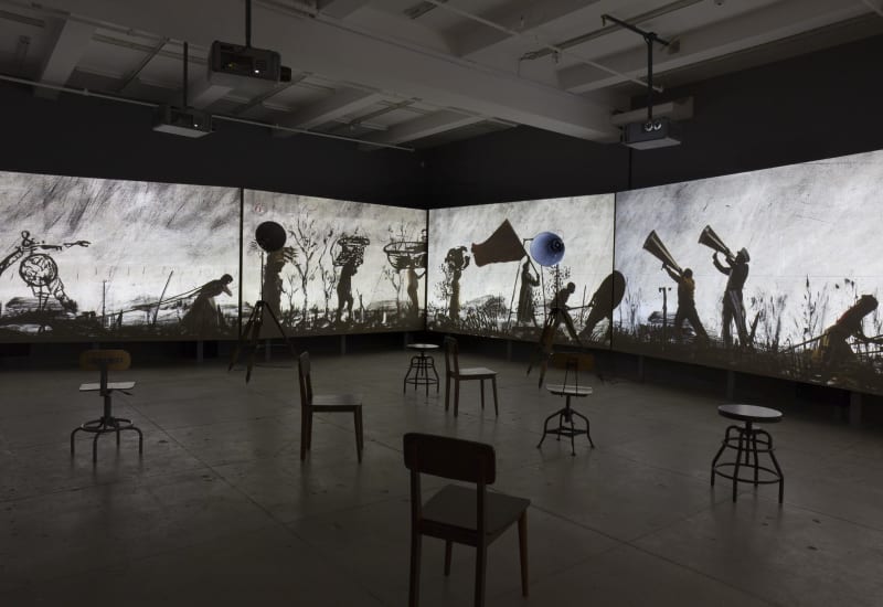 A large darkroom with scattered chairs is illuminated by projections of silhouetted figures.