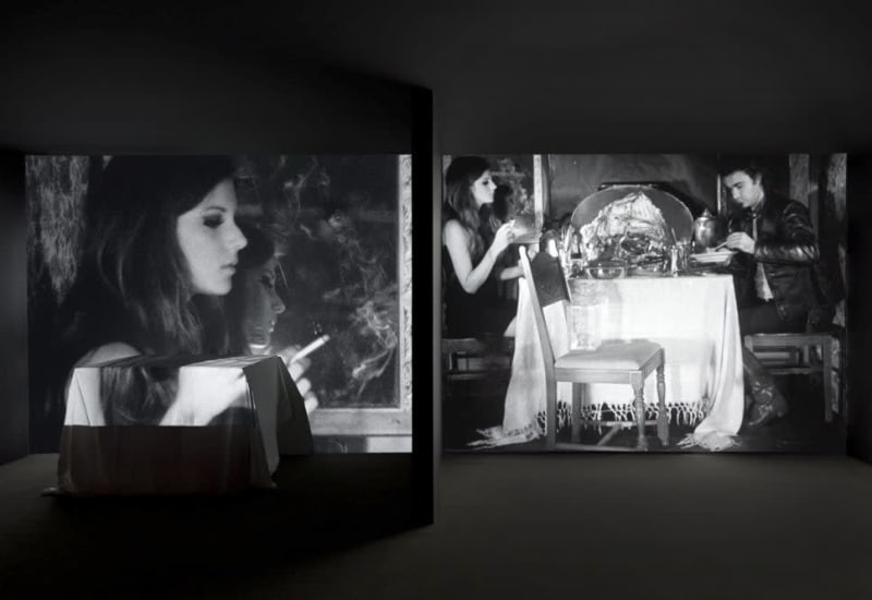 A dark room is illuminated by a projected diptych of a black and white image of a woman and man at a dining table. 