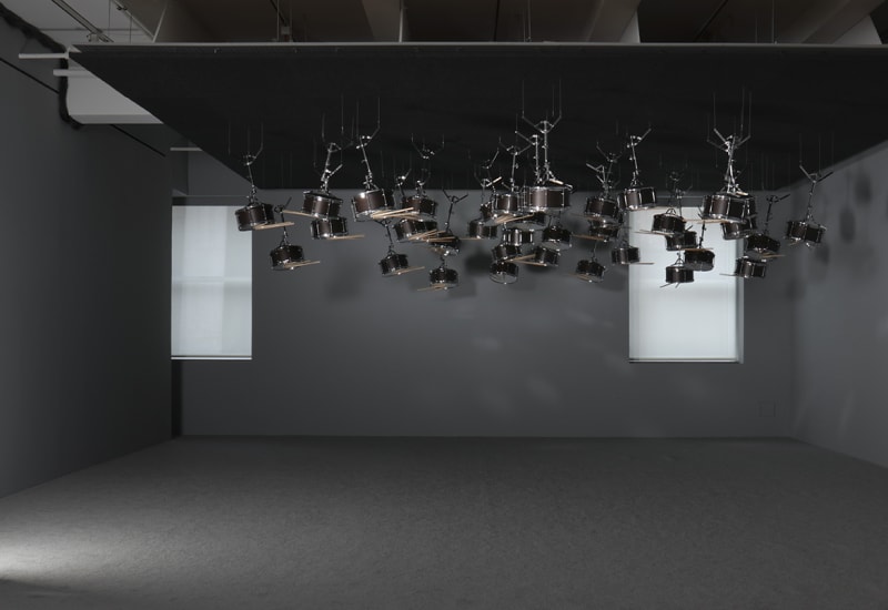 Installation shot of many snare drums and drumsticks hanging upside down from a black drop ceiling.