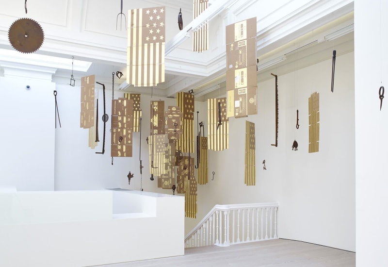 Gallery view of numerous cardboard flags and various tools suspended from ceiling.